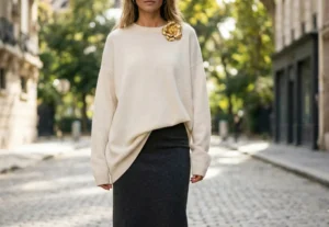 A woman stands on a cobblestone street, wearing a cream-colored oversized knit sweater with a large gold floral brooch and a dark grey midi skirt. The background shows blurred European-style buildings.