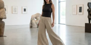 A woman wearing wide-leg beige linen trousers and a black sleeveless top stands confidently in a spacious, minimalist art gallery surrounded by abstract sculptures.