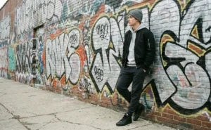 A young man in a black bomber jacket, hoodie, and beanie leans against a brick wall covered in colorful graffiti.