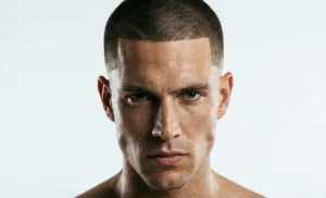 A portrait of a bearded man with a modern comb-over haircut and faded sides, holding a coffee cup and looking out a window in a brick-walled cafe.