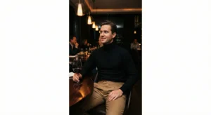 "A man seated at a table in a dimly lit, upscale restaurant. He is wearing a black fitted turtleneck tucked into tan high-waisted Gurkha trousers and holding a glass of red wine. The lighting is warm and ambient."