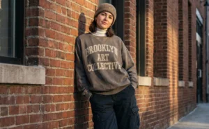 A portrait of a young woman leaning against a red brick wall, enjoying the afternoon sun. She wears a brown beanie, a grey "BROOKLYN ART COLLECTIVE" sweatshirt layered over a white turtleneck, and dark cargo pants with hands in her pockets.