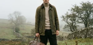 A man standing in a foggy English countryside landscape, wearing an olive green waxed cotton jacket, a beige turtleneck sweater, and dark trousers. He is holding a large brown leather weekend bag. The atmosphere is misty with muted earth tones and stone walls in the background.