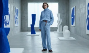 A full-body photograph of a woman standing in a minimalist, concrete art gallery. She is wearing a monochrome outfit consisting of a chunky oversized powder blue turtleneck sweater and matching wide-leg trousers, cinched with a brown belt. She is surrounded by large abstract blue and white sculptures.
