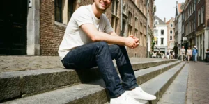 A natural photograph of a man smiling while sitting on stone steps outdoors. He is wearing a light-colored t-shirt, dark raw denim jeans, and clean white leather sneakers.