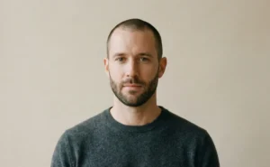 A minimalist studio portrait of a man with a buzz cut and a very short, neatly groomed beard, wearing a dark grey cashmere sweater against a plain neutral background.