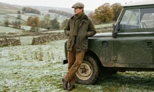 A man in the countryside wearing a flat cap, green field jacket, and brown corduroy trousers, leaning against a vintage Land Rover.