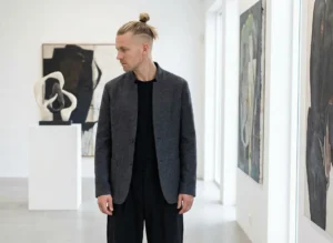 A man standing in a modern art gallery, with his blonde hair tied up in a high top knot bun and shaved undercut sides.