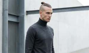 A sleek fashion photograph of a man with shaved sides and a small, neat top knot bun, wearing a dark grey turtleneck and standing against a minimalist concrete architectural wall.