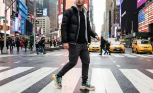 A man crosses a busy city street in a crosswalk, wearing a layered streetwear outfit with a black puffer jacket, grey hoodie, and cargo joggers.