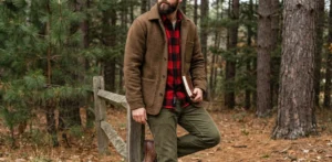A bearded man in a brown wool jacket and red plaid shirt leans against a wooden fence in a forest.