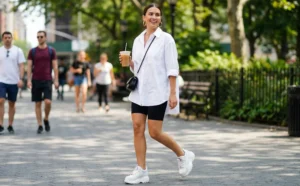 A smiling woman walking on a city sidewalk holding an iced coffee, wearing an oversized white shirt, black bike shorts, and white sneakers.