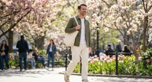 A candid shot of a smiling man walking through a sunny city park during spring. He wears an olive green overshirt over a white t-shirt and ivory denim jeans, carrying a canvas tote bag. Blooming cherry blossom trees and colorful tulips are visible in the background.