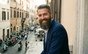 A man with a full, well-groomed brown beard smiles happily while standing on a balcony overlooking a European street during the day.