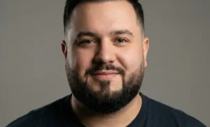 A front-facing portrait of a man with a round face, featuring a beard with a high fade on the cheeks and an angular shape at the chin.