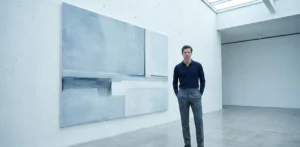 A man wearing slate grey wool trousers and a navy blue long-sleeve polo shirt, standing in a minimalist art gallery next to a large abstract painting in blue and grey tones.