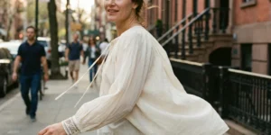 A candid shot of a smiling woman walking down a city sidewalk, looking back over her shoulder. She wears a loose, cream-colored peasant blouse with embroidered cuffs that billows in the wind.