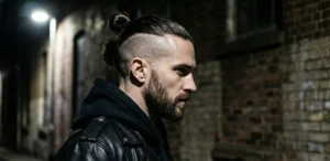 A profile view of a man in a black leather jacket and hoodie at night. He has a man bun with shaved undercut sides and a full beard, standing in a dark alleyway with a brick wall.
