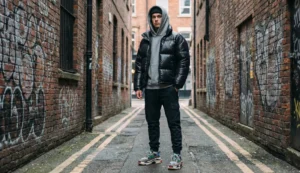 A young man wears a black glossy puffer jacket over a grey hoodie in a graffiti-covered brick alleyway.