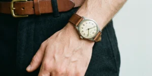 A detailed close-up of a man's wrist wearing a simple vintage-style analog watch with a brown leather strap. His hand is resting near a brown belt on dark trousers.