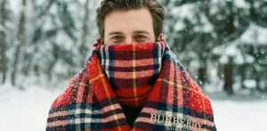 A portrait of a man outdoors in the snow, with his lower face wrapped in a thick, red tartan plaid Burberry scarf showing the embroidered logo, against a snowy forest background.