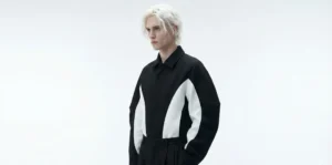 A high-fashion studio photograph of a male model with loosely styled, near-white platinum blonde hair. He is wearing a structured, black and white avant-garde jacket and black trousers against a plain, bright white background.