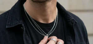 A close-up detail of a man's neck and chest area. He is wearing a black t-shirt under a black denim jacket, adorned with three layered silver minimalist chains. His hand, wearing two silver rings, is holding the necklaces.