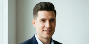 A polished portrait of a smiling man with a square face shape, featuring a neatly tapered quiff haircut with a smooth transition to shorter sides, wearing a navy blazer.