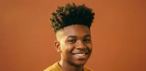 A studio headshot of a smiling Black teenage boy with a high skin fade haircut and voluminous natural curls on top.