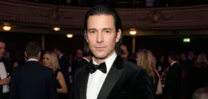 A man in a black velvet tuxedo and bow tie, with his shoulder-length hair expertly slicked back behind his ears. He is standing in a dimly lit, ornate event hall.