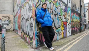A young man dressed in trendy streetwear, including a bright blue oversized puffer jacket and a beanie, leaning against a graffiti-covered wall in an alley.