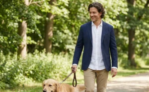 A smiling man with wavy hair, wearing a navy blazer and chinos, walks a golden retriever dog on a leash along a path in a green park.