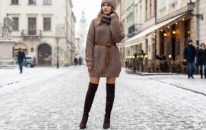 A woman posing on a snowy cobblestone street, wearing a chunky brown knit sweater dress, a matching beanie, and tall suede boots.