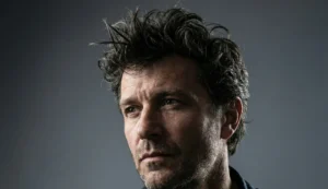 A dramatic portrait of a man with dark, thick, textured hair styled into a messy, high-volume quiff, looking off-camera against a dark grey background with side lighting.