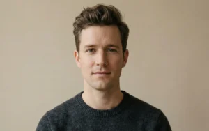 A minimal portrait of a man with a casual, natural quiff hairstyle wearing a dark grey crewneck sweater against a plain neutral beige background.