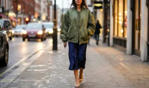 A woman walks on a city sidewalk at dusk, wearing an olive green satin bomber jacket, a navy blue satin midi skirt, and gold strappy heels. The background shows blurred car lights and illuminated shop windows.