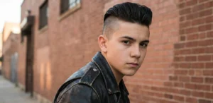 A portrait of a serious teenage boy in a black leather jacket, leaning against a brick wall. He has a disconnected undercut hairstyle with dark hair slicked high on top and sharply shaved sides.