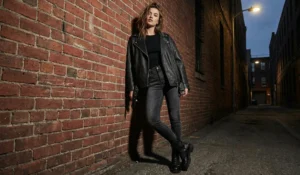 An edgy fashion shot of a young woman leaning against a textured brick wall in a dimly lit urban alley at dusk. She wears an oversized black leather moto jacket over a black top, dark skinny jeans, and chunky combat boots. A streetlamp illuminates her from above.