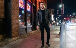 Man wearing a black leather jacket and black jeans with boots standing outside, showing an edgy classic style.