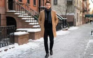 Man walking on a snowy street wearing a camel coat and black jeans, a sharp winter outfit for men.