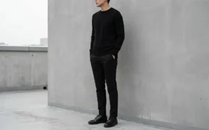 Man standing against a grey wall wearing an all-black outfit with a sweater and trousers for sleek minimal style.