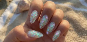 A close-up sunlit view of long, translucent almond-shaped nails with encapsulated chunky iridescent glitter flakes shifting in colors of aqua, lilac, and seafoam green.