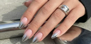 A trendy shot of almond-shaped nails displaying a sheer pink base that fades into bold, highly reflective silver mirror-chrome gradient tips. The hand wears a chunky silver ring and rests on concrete near metallic objects.