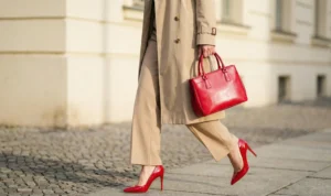 A close-up view of a woman walking on cobblestones, highlighting the contrast of her bright cherry red stiletto heels and matching red leather handbag against her neutral beige trench coat and trousers.
