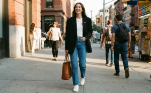 A stylish woman smiling broadly while walking down a sunny city sidewalk, wearing an oversized black blazer, jeans, and white sneakers, and carrying a brown leather tote bag.