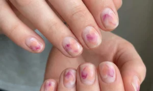 Close-up view of short, natural nails featuring a hazy, watercolor-style design. Patches of magenta and peach ink blur into indistinct, melting heart shapes on a sheer, milky base with a soft matte finish.