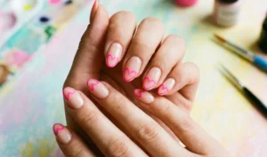 A playful photograph of hands with oval nails and a clean nude base. The tips are defined by fine neon pink outline drawings of heart shapes, with the heart's cleft at the center edge.