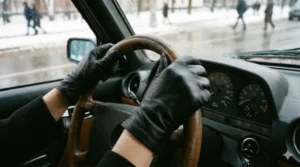 A close-up view from inside a car, showing hands wearing sleek black leather gloves gripping a wooden steering wheel on a snowy day.