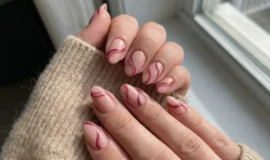 Close-up of oval nails with a glossy nude base, decorated with unique abstract wavy lines in berry and deep pink hues, resting on a beige knit sweater.