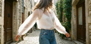 A woman in a sheer white blouse and blue jeans spins joyfully, arms outstretched, in a sun-drenched cobblestone alleyway lined with old stone buildings.